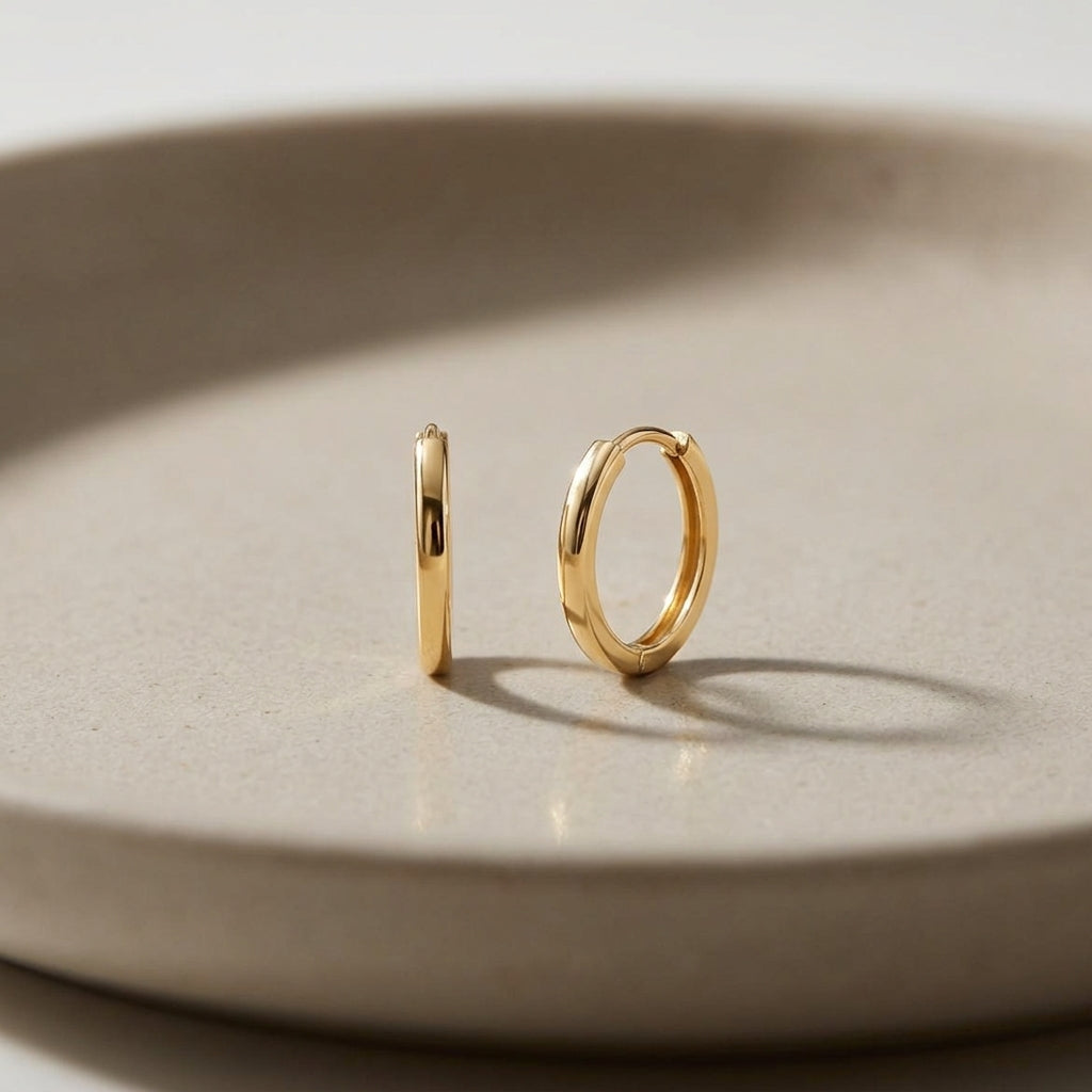Pair of 14k gold huggie hoop earrings, 15mm, displayed on a neutral matte ceramic, emphasizing the minimalist design and polished finish.